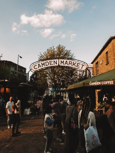 The image depicts the entrance to Camden Market, with a prominent arched sign that reads 'CAMDEN MARKET' positioned above the street. The scene is set during daytime with partly cloudy skies visible a