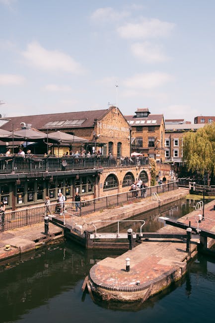 A daytime view of a canal-side building with a brick facade and arched windows, featuring an outdoor terrace with dark metal railings and several tables and chairs with people seated, some under umbrellas. The terrace overlooks the canal, which has a small round dock or extension with mooring posts and white bollards. A metal ladder is attached to the dock, and a concrete walkway runs alongside the water’s edge. In the background, additional buildings with brick and modern facades are visible, along with trees and a bright blue sky with scattered clouds. The scene captures a typical urban waterside setting suitable for home relocation or furniture transport planning, with visible elements such as packed boxes, moving equipment, or vehicles not present in this specific image, but suggesting a location associated with relocations and moving logistics, consistent with services offered by Removal Companies Camden.