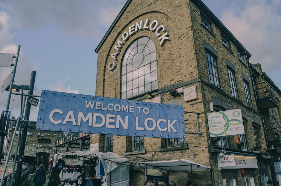 A three-story brick building with large arched windows featuring black frames, located on Camden Lock in Camden Town. The building has a prominent sign at the top reading 'CAM DEN LOCK,' and a large blue banner hanging in front that displays 'WELCOME TO CAMDEN LOK' in bold white letters. Surrounding the building, there are various signs, including one indicating 'Camden Lock & Shops to Rent' with a phone number, and a smaller sign for a supermarket. In the foreground, there is a covered outdoor area with a white tent or canopy, beneath which retail stalls are visible, displaying clothing and accessories. Nearby, pedestrians can be seen walking along the pavement, and parked vehicles are present adjacent to the building. The scene is lit by natural daylight, with some cloud cover in the sky above. The image reflects an active marketplace environment, typical of a busy area involved in home relocation and furniture transport services, as organised by Removal Companies Camden.