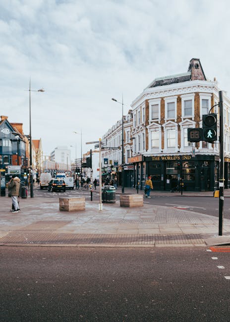 A busy street scene in Camden featuring a pedestrian crossing with a green walking signal, surrounded by mixed-use buildings including a prominent corner property with decorative architectural details and a sign reading 'The World's End.' The scene includes several pedestrians waiting or walking, some carrying bags, and vehicles such as cars and vans moving or parked along the street. Two large wooden crates are placed on the pavement near the crossing, and behind them, a person can be seen lifting or positioning a cardboard box. The area appears to be part of a home relocation process with a van parked nearby for furniture transport or packing and moving activities, consistent with professional removals services by Camden-based companies. The overcast sky provides diffused natural lighting, illuminating the urban environment which emphasizes the logistics and movement typical during property moves in Camden.