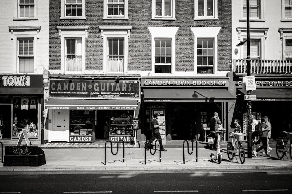 A black and white photograph of a busy street frontage in Camden, showing three small retail shops situated on the ground floor of a multi-storey brick building. The shops include a music store with a sign reading 'CAMDEN GUITARS', a bookshop named 'CAMDEN TOWN BOOKSHOP', and an additional shop partially visible to the right. The pavement in front of the shops features several pedestrians walking, some carrying bags or backpacks, and one person using a bicycle. Bicycles are parked along the street, secured to bike racks. In front of the shops, a few cardboard boxes and packing materials are visible, along with a person lifting a box onto a trolley for transport, indicating a moving or delivery process. The scene includes street fixtures such as a lamp post, a parking sign, and a bike rack, with the outdoor lighting suggesting daytime conditions. This image illustrates the logistical aspects of home relocation or furniture transport, as practiced by Removals companies Camden, with items prepared for moving or delivery outside the shop.