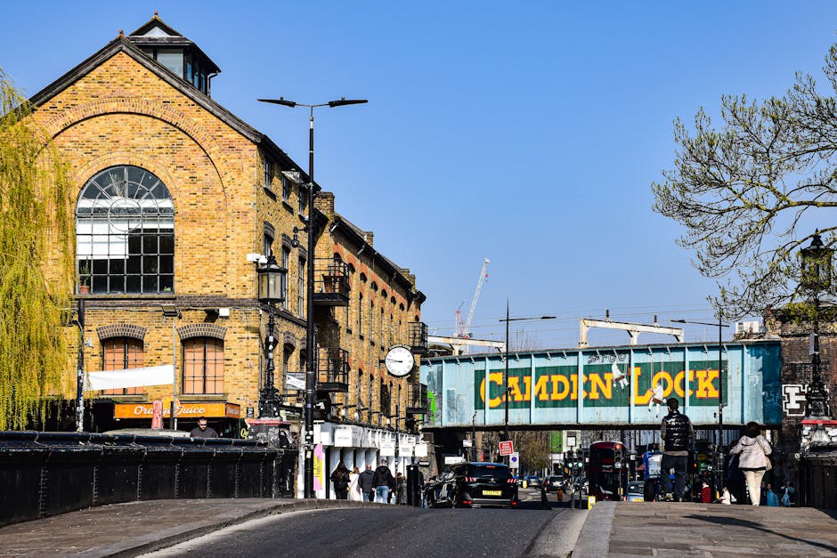 A view of Mornington Crescent in Camden, featuring a brick building with large arched windows and a pitched roof, situated along a busy street with pedestrians walking on the pavement. In the background, there is a blue railway bridge with the words 'Camden Lock' painted in yellow, and a crane can be seen in the distance. On the street, several cars are parked or moving, and a black van is partially visible near the building. Trees with light foliage line the street, and street lamps and clocks are mounted on lampposts along the sidewalk. The scene depicts an urban area commonly associated with home relocation and moving logistics, with visible elements like street furniture and transportation infrastructure relevant to furniture transport and loading processes. The overall environment is well-lit under clear weather, emphasizing the mix of residential and commercial activity in Camden.
