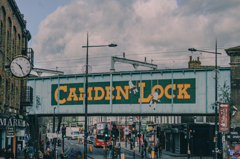 A black and white photograph of a busy street frontage in Camden, showing three small retail shops situated on the ground floor of a multi-storey brick building. The shops include a music store with a sign reading 'CAMDEN GUITARS', a bookshop named 'CAMDEN TOWN BOOKSHOP', and an additional shop partially visible to the right. The pavement in front of the shops features several pedestrians walking, some carrying bags or backpacks, and one person using a bicycle. Bicycles are parked along the street, secured to bike racks. In front of the shops, a few cardboard boxes and packing materials are visible, along with a person lifting a box onto a trolley for transport, indicating a moving or delivery process. The scene includes street fixtures such as a lamp post, a parking sign, and a bike rack, with the outdoor lighting suggesting daytime conditions. This image illustrates the logistical aspects of home relocation or furniture transport, as practiced by Removals companies Camden, with items prepared for moving or delivery outside the shop.